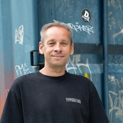 Joost Faassen in een zwart t-shirt, glimlachend, voor een blauwe stalen deur vol graffiti.